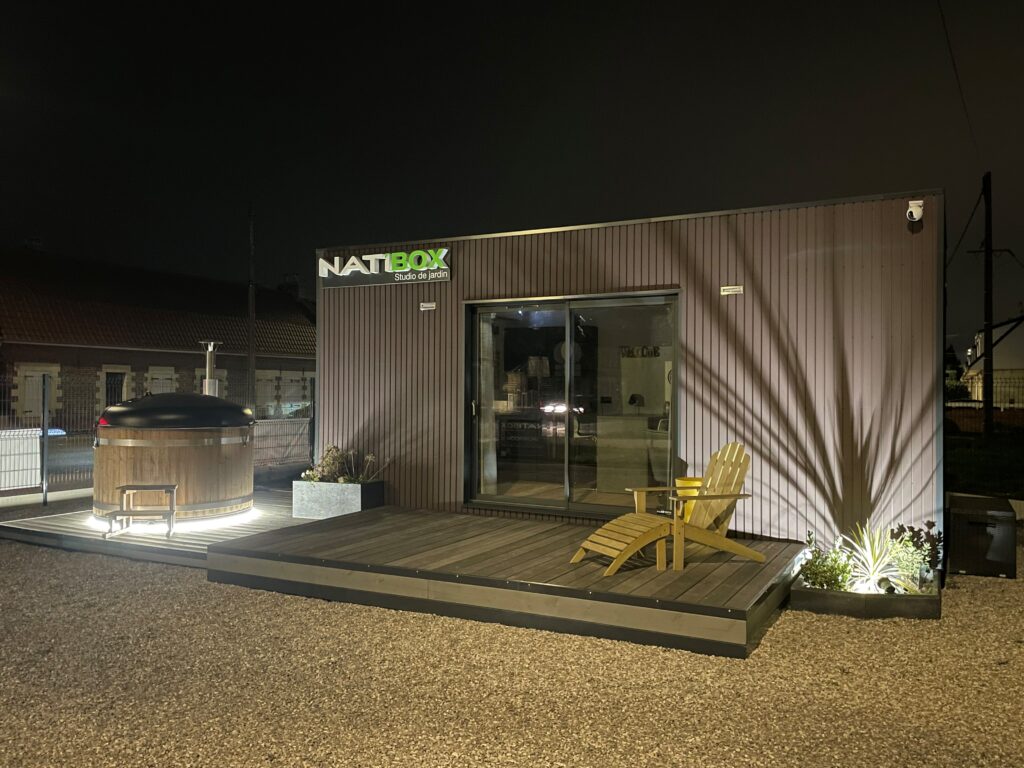 Terrasse en bois modulaire de nuit avec bain nordique éclairé et fauteuil en bois devant un module Natibox.